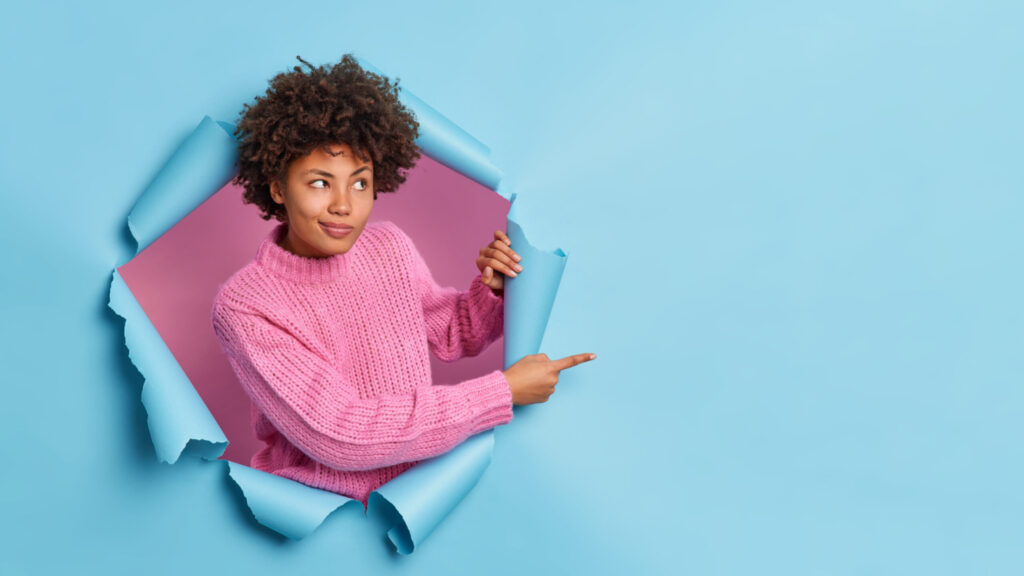 curly haired young woman gives advice where to go indicates place for advertisement breaks through blue wall wears knitted sweater recommends something scaled e1683252407864